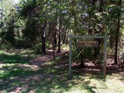 Sign for Meadowbrook Trail at the entrance of a wooded path, surrounded by trees and greenery.