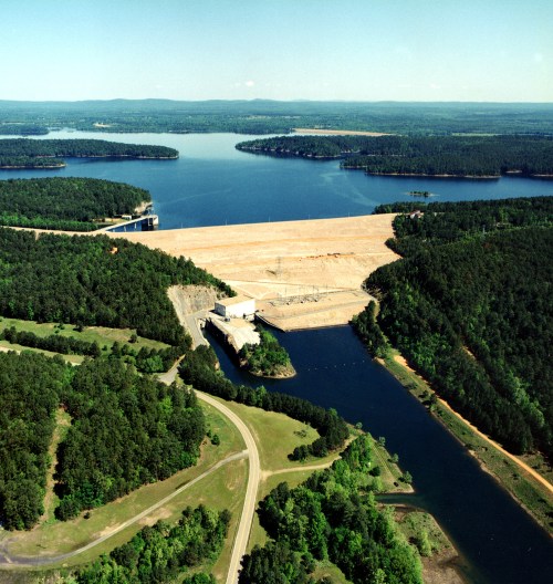 Aerial view of a large reservoir surrounded by forested land and a dam, with calm water and distant hills.