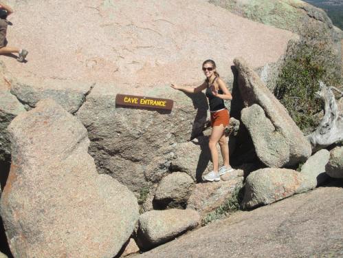 A woman in athletic wear stands next to a sign that reads "Cave Entrance" on a rocky surface.
