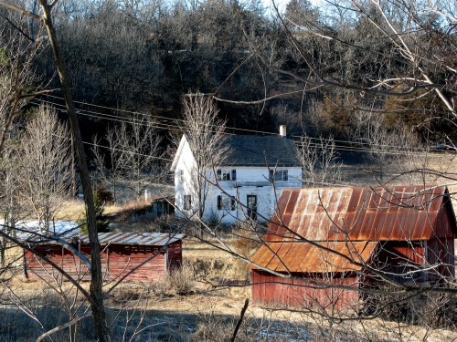A white farmhouse and red barns nestled in a rural landscape, surrounded by bare trees and rolling hills.