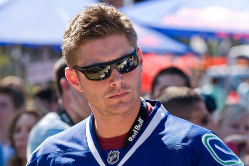 A man wearing sunglasses and a blue sports jersey stands outdoors, surrounded by a crowd.