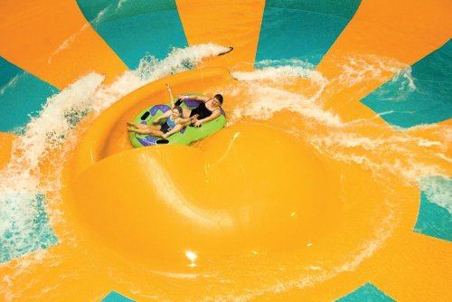 Two people enjoy a water slide, riding a green and blue inner tube on a vibrant orange and teal surface.
