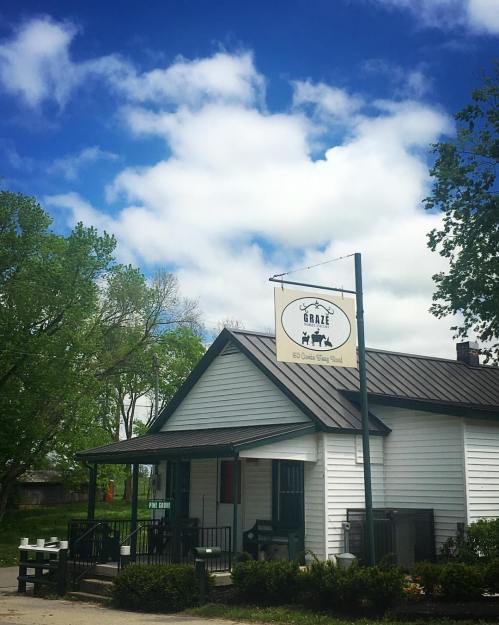 A small white building with a sign reading "Graze" under a blue sky with clouds, surrounded by green trees.