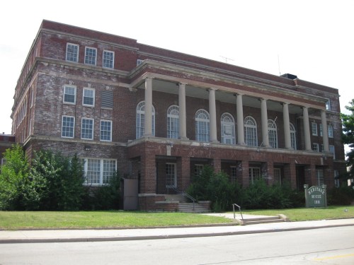 Historic brick building with columns and large windows, surrounded by greenery and a grassy area.