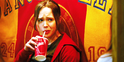 A person in a red hoodie sips from a cup, looking slightly confused or concerned, with colorful banners in the background.