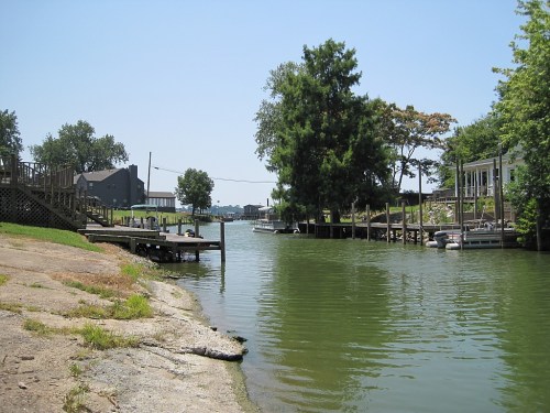 A serene waterway lined with trees and houses, featuring docks and boats under a clear blue sky.