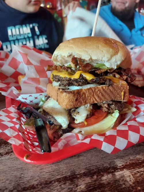 A towering burger with layers of meat, cheese, bacon, lettuce, and tomato, served in a red basket with checkered paper.