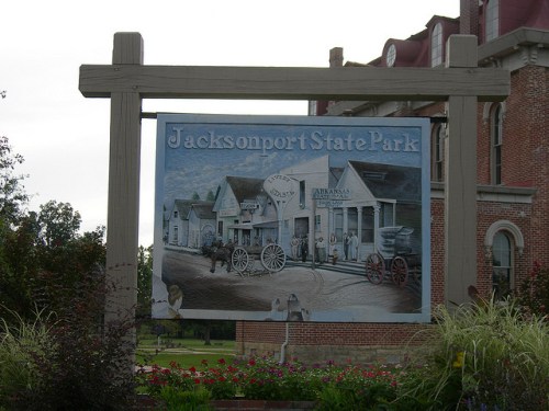 Sign for Jacksonport State Park featuring a mural of historic buildings and wagons, surrounded by greenery.