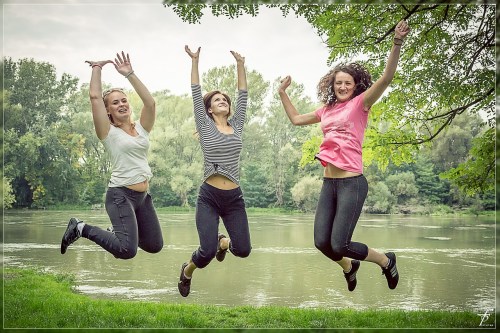 Three women joyfully jumping in the air by a serene river, surrounded by lush greenery.