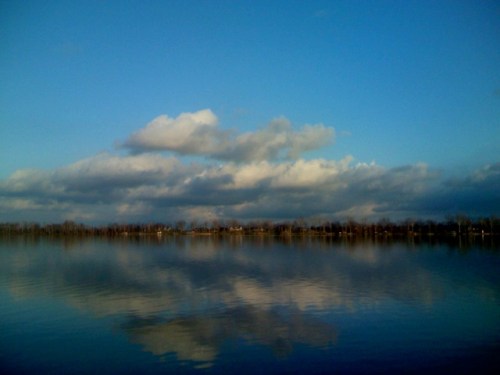 A serene lake reflecting fluffy clouds and a clear blue sky, surrounded by trees along the shoreline.