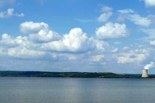 A serene lake scene with fluffy clouds and a cooling tower in the distance, surrounded by green hills.