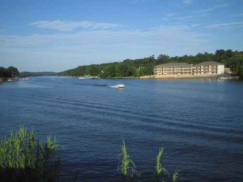 A serene river scene with a boat gliding past lush green banks and a building in the background under a clear blue sky.