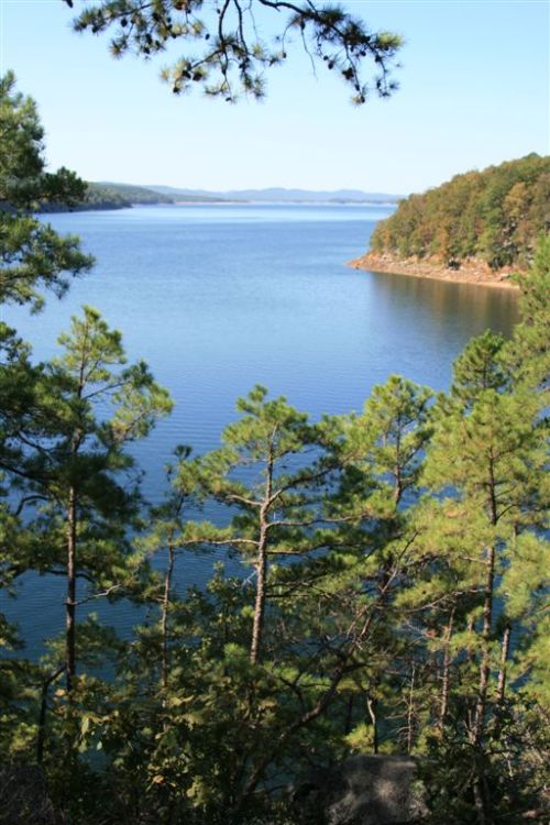 A serene lake surrounded by trees, with distant hills under a clear blue sky.