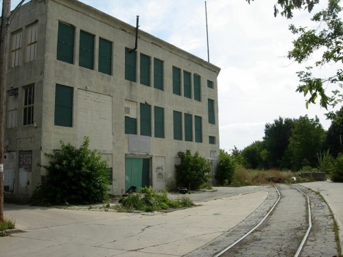 Abandoned building with boarded windows, overgrown vegetation, and a railway track beside it under a cloudy sky.