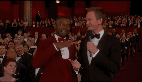 Two men in formal attire smile and interact on stage at an awards ceremony, with an audience visible in the background.