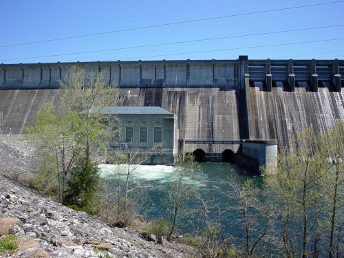 A large concrete dam with a power station beside a flowing river, surrounded by greenery under a clear blue sky.