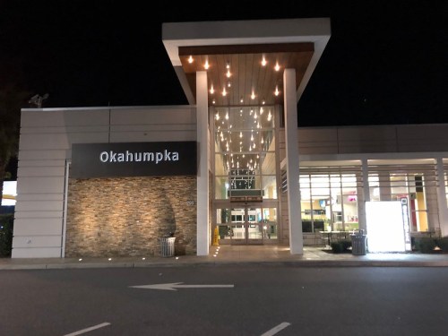 Night view of a modern building with "Okahumpka" sign, featuring large glass doors and bright interior lights.