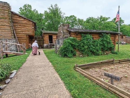 A woman in historical attire walks along a path by log cabins and a tobacco garden, with trees and an American flag nearby.