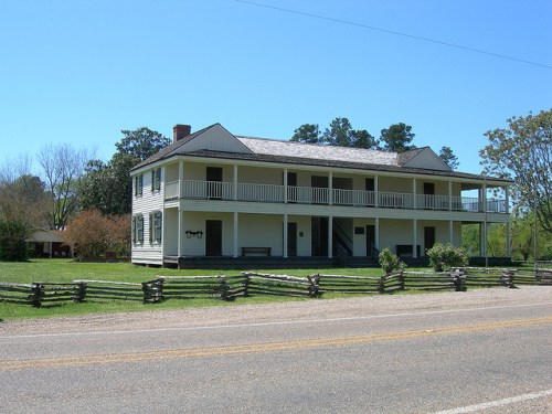 A two-story yellow house with a porch, surrounded by greenery and a wooden fence, set against a clear blue sky.
