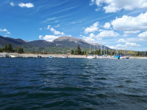 A serene lake scene with boats docked along a sandy shore, surrounded by mountains and a blue sky with fluffy clouds.