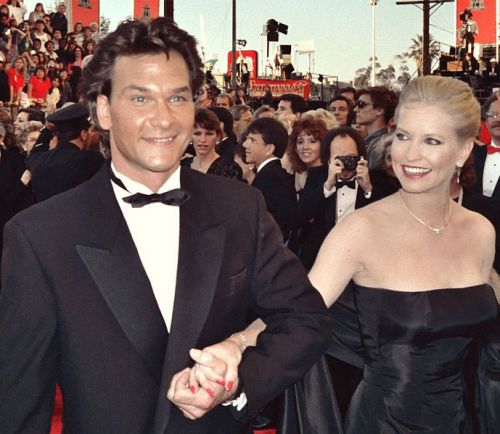 A man in a tuxedo and a woman in a black gown walk hand in hand at a red carpet event, surrounded by a crowd.