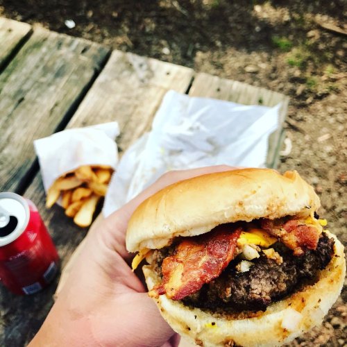 A hand holding a bacon cheeseburger with fries and a soda in the background on a wooden surface.