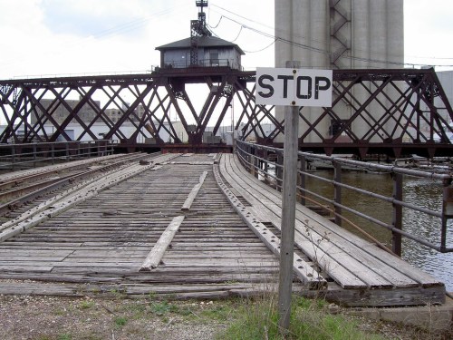 A wooden bridge with a "STOP" sign, leading to a raised structure over water, surrounded by industrial scenery.