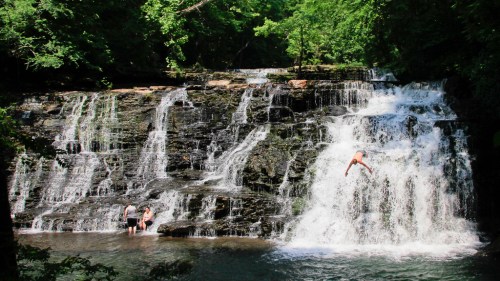 A scenic waterfall with people swimming and one person jumping into the water, surrounded by lush greenery.