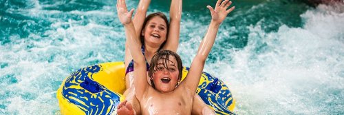 Two children joyfully ride a yellow inflatable tube down a water slide, splashing in the water.