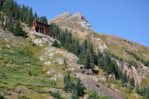 A weathered wooden structure on a mountainside, surrounded by evergreen trees and rocky terrain under a clear blue sky.