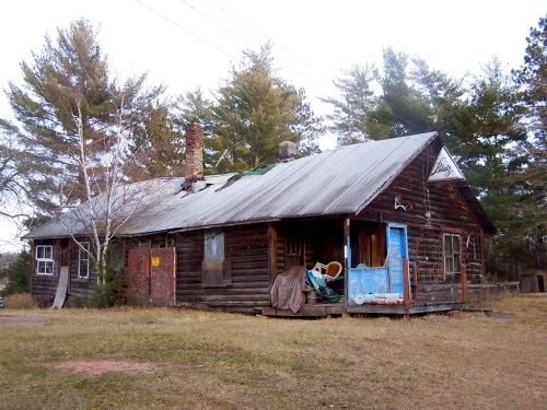 An old, weathered wooden cabin with a metal roof, surrounded by trees and overgrown grass.