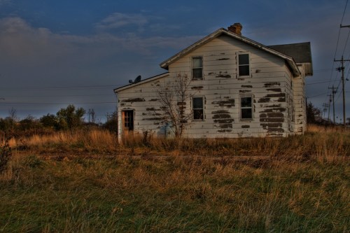 Abandoned white house with peeling paint, surrounded by tall grass and power lines under a cloudy sky.