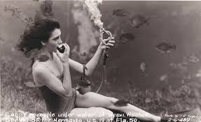 A woman in a vintage swimsuit uses a phone underwater, surrounded by fish, in a black-and-white photo.