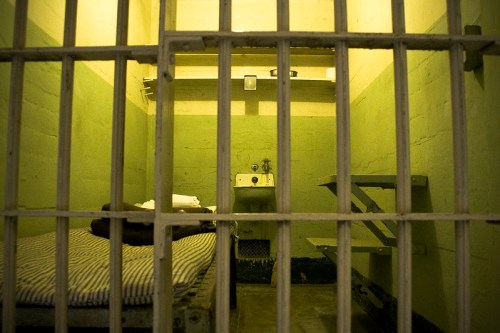 A dimly lit prison cell with a bed, sink, and barred window, conveying a sense of confinement.