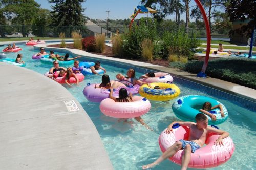 A group of people relaxes on colorful inner tubes in a sunny pool, surrounded by greenery and play structures.