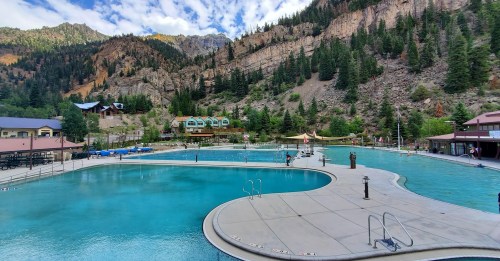 A large outdoor pool surrounded by mountains and trees, with people enjoying the water on a sunny day.