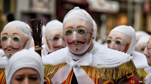 A group of people in traditional costumes and masks, featuring white fabric and green glasses, parading in a festive setting.