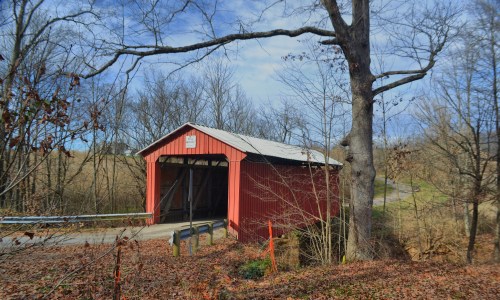 A red covered bridge surrounded by bare trees and autumn leaves, set against a clear blue sky.