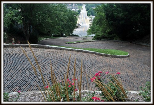 A winding cobblestone path lined with greenery and flowers, leading to a distant road and trees in the background.