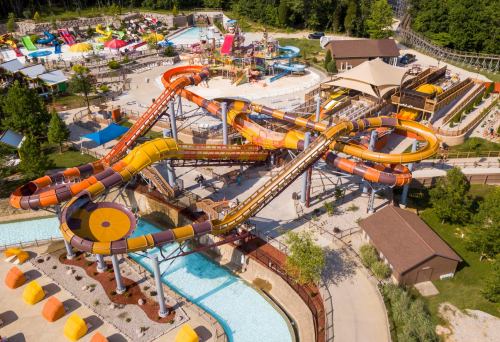 Aerial view of a colorful water park featuring multiple slides, pools, and lounging areas surrounded by trees.