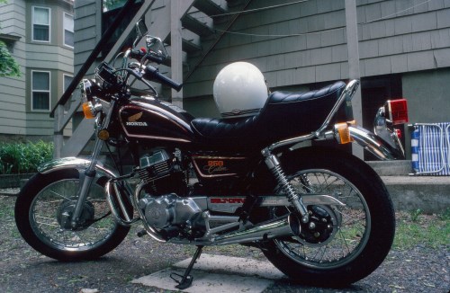 A black Honda motorcycle parked on gravel, with a white helmet resting on the seat, near a house and stairs.