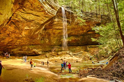 A scenic waterfall cascades from a rocky cliff, with people exploring the sandy area below in a lush forest setting.