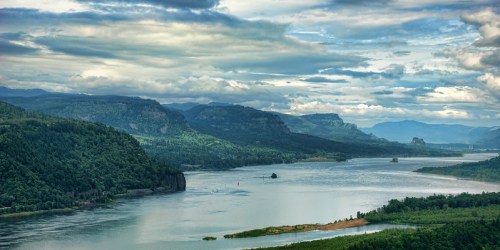 A serene river flows through lush green mountains under a cloudy sky, creating a tranquil landscape.