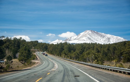 A winding road leads through green trees, with a snow-capped mountain in the background under a clear blue sky.