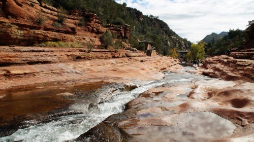 A serene river flows through red rock formations, surrounded by greenery and mountains under a cloudy sky.