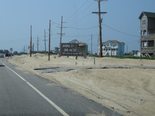 Sign for Mirlo Beach along a sandy road, with beach houses and power lines in the background under a clear sky.