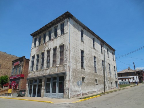 Abandoned three-story building with peeling paint, large windows, and a sloped street under a clear blue sky.