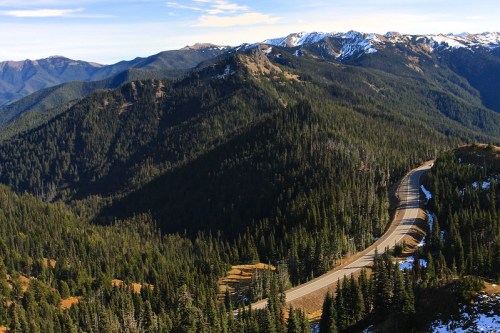 A winding road cuts through a mountainous landscape, surrounded by dense evergreen forests and snow-capped peaks.