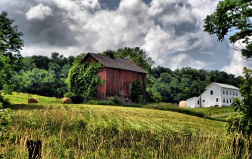 A red barn and a white house surrounded by green fields and trees under a cloudy sky. Hay bales are scattered nearby.