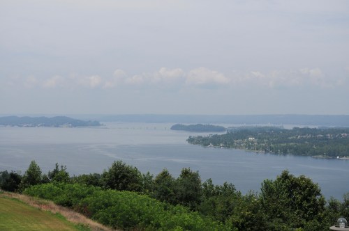 A scenic view of a calm lake surrounded by green trees and distant hills under a cloudy sky.